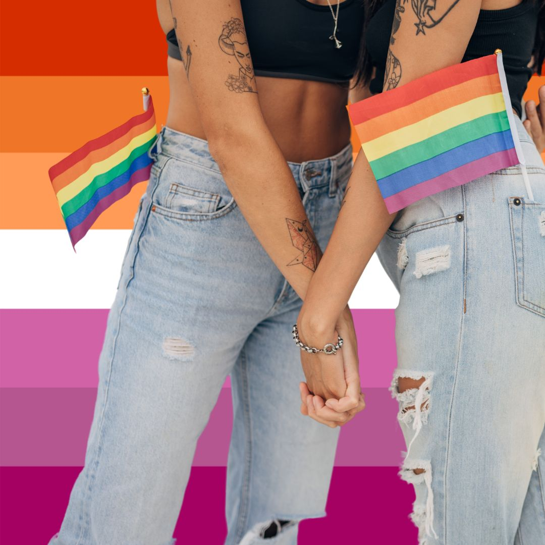 Two girls holding hands with small gay pride flags in their back pockets with tattoos in their arms standing in front of the lesbian flag with the word lesbian below them