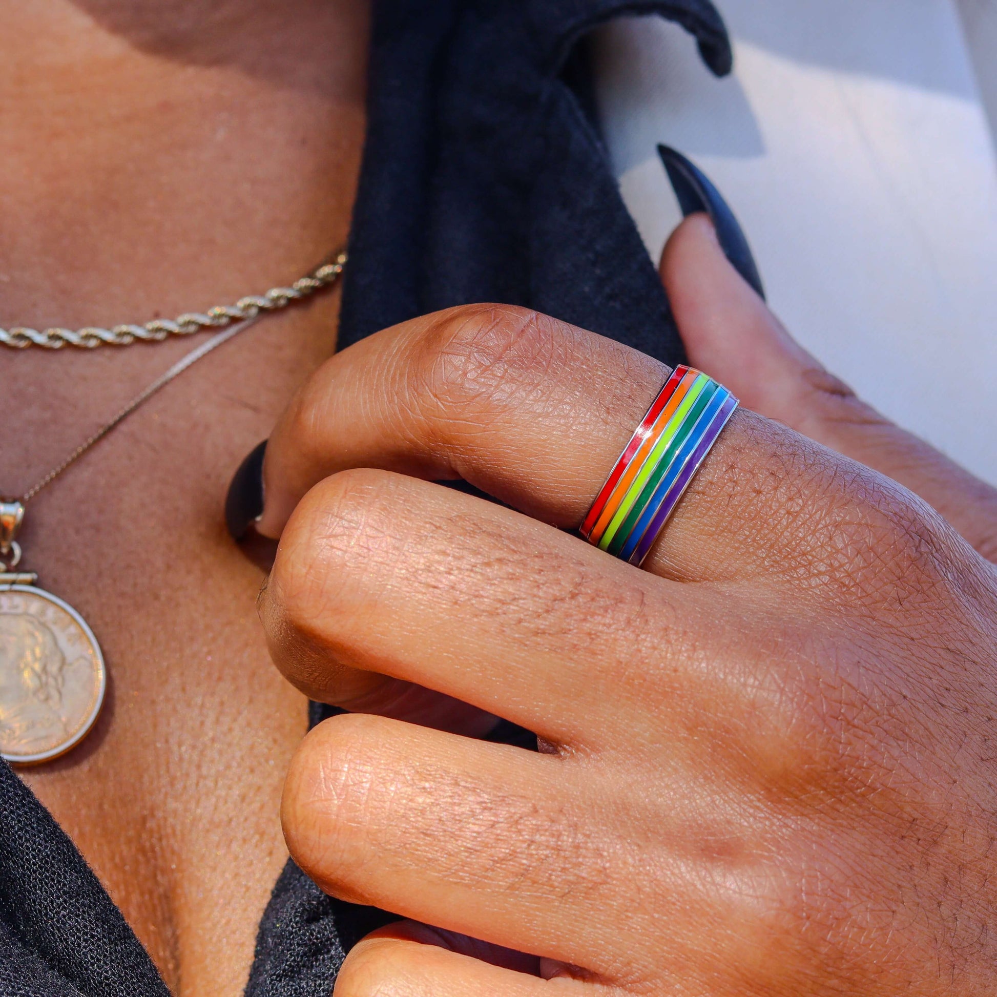 Rose Gold Co Rainbow Pride Ring on index finger of a brown skinned woman with black fingernails and an open collar