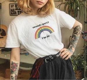 A young woman with short blonde hair and glasses wearing a white t-shirt with the words "Sounds Gay I'm In" in rainbow